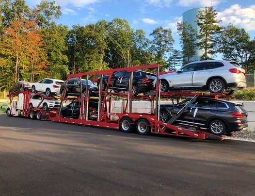 Open car carrier loaded with BMW and Porsche SUVs departing for highway delivery — Baier Transport door-to-door auto shipping service